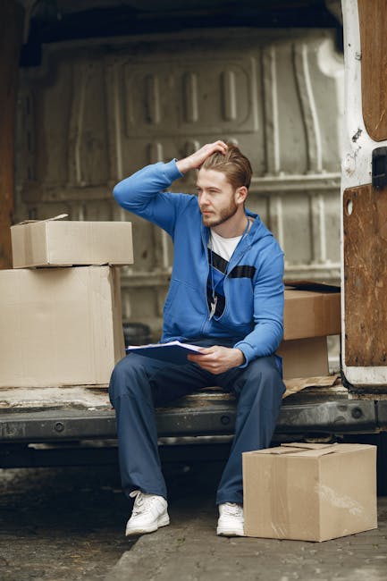 A young man with short, light brown hair and a beard is sitting on the edge of an open moving van in a garage or driveway area, surrounded by cardboard boxes of various sizes that are either stacked o