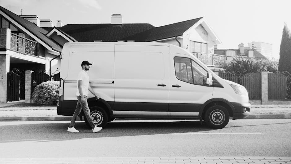 A young man with short, light brown hair and a beard is sitting on the edge of an open moving van in a garage or driveway area, surrounded by cardboard boxes of various sizes that are either stacked o