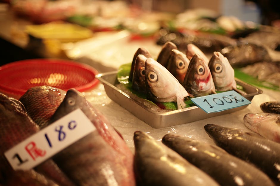 Close-up image of a fish market display showing fresh fish and seafood arranged on a bed of crushed ice. In the foreground, there are whole fish with shiny scales and a label indicating their price at 16.80. Behind them, a metal tray holds several fish heads with visible eyes and gills, arranged upright on a bed of green leaves, with a small sign marked 10.00. To the left, additional fish are partially visible, some wrapped in clear plastic or placed on ice, with other seafood items in the blurry background, suggesting a busy market environment. The scene is well-lit, highlighting the textures of the fish and seafood. This image relates to food delivery or seafood sales, which may connect to the logistics involved in house removals when transporting perishable goods, and is situated in an indoor setting typical of fish markets.