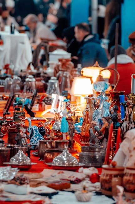 A variety of vintage and decorative household items displayed on a table at an indoor market or auction, including glass and ceramic figurines, ornate vases, lamps, and metallic decorative objects. The items are arranged closely together on tablecloths with some items partially obscured by others. In the background, blurred figures of people browsing and examining items can be seen, suggesting a busy environment typical of house clearance or moving situations. The lighting is warm, highlighting the details of the collectibles, which are often packed with cushioning materials like tissue paper or fabric. This scene exemplifies the types of contents that can be encountered during house removals, with [COMPANY_NAME] involved in packing, transporting, or relocating such items as part of their house or commercial removal services. The setting appears to be an auction hall or a market dedicated to furnishings, antiques, or collectibles, coordinated in a way that is consistent with professional removals' handling of fragile and valuable objects.