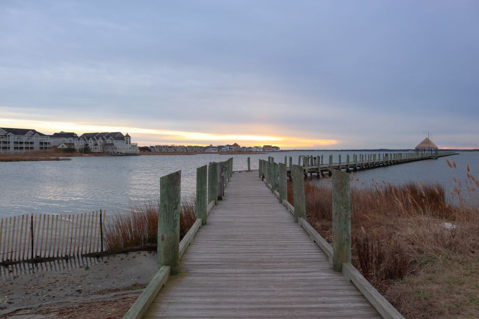 A wooden pier extends from the foreground into the water, supported by greenish wooden posts on either side. The pier appears to be part of a house removal or relocation project, with the scene set on a calm waterfront area during sunset or early evening. Surrounding the pier are dry grasses and a sandy shoreline, with a weathered wooden fence partially visible on the left side. In the background, there are modern residential buildings along the water's edge, and the sky is mostly cloudy with a hint of sunlight near the horizon, creating a serene and structured environment suitable for loading or unloading furniture and moving equipment. The image highlights the outdoor environment typical of house removals in Nags Head, where careful logistics are essential for smooth furniture transport and home relocation processes, supported by professional services like Man with Van Nags Head.