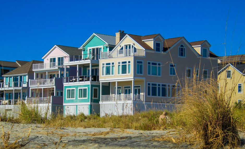A wooden pier extends from the foreground into the water, supported by greenish wooden posts on either side. The pier appears to be part of a house removal or relocation project, with the scene set on a calm waterfront area during sunset or early evening. Surrounding the pier are dry grasses and a sandy shoreline, with a weathered wooden fence partially visible on the left side. In the background, there are modern residential buildings along the water's edge, and the sky is mostly cloudy with a hint of sunlight near the horizon, creating a serene and structured environment suitable for loading or unloading furniture and moving equipment. The image highlights the outdoor environment typical of house removals in Nags Head, where careful logistics are essential for smooth furniture transport and home relocation processes, supported by professional services like Man with Van Nags Head.