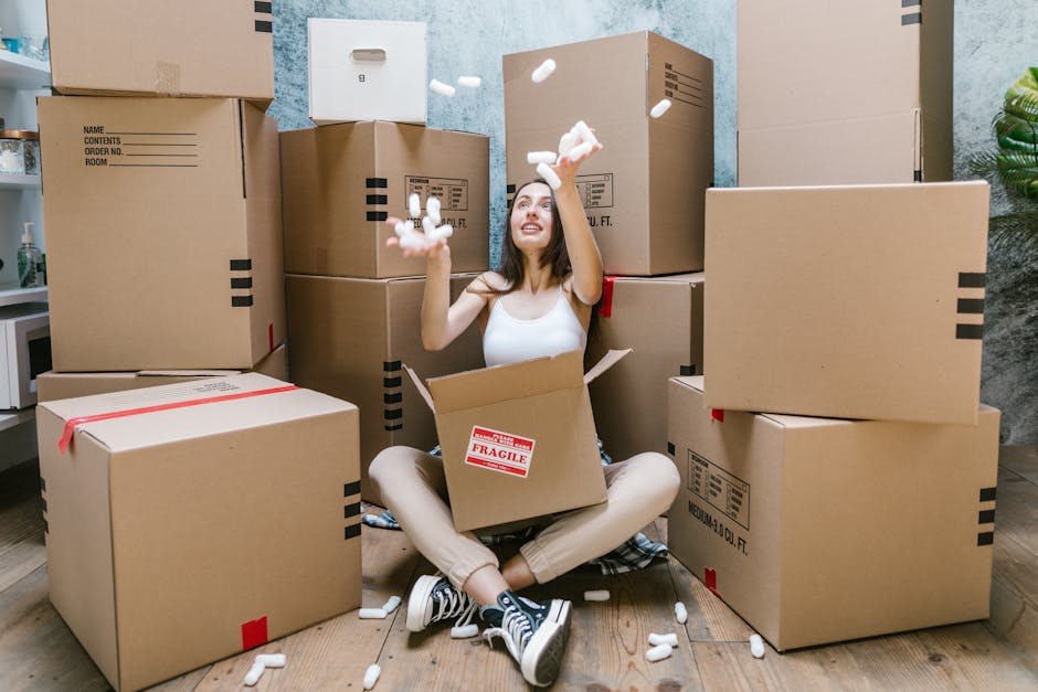 A young woman with long dark hair, sitting cross-legged on a wooden floor inside a room, is surrounded by numerous packed cardboard boxes of various sizes. She is wearing a white sleeveless top, beige pants, and black-and-white striped sneakers. She is smiling and looking up while tossing packing foam peanuts into the air, with some falling into an open box placed on her lap. The boxes are sealed with clear or red tape, and some have labeling or handling stickers, such as a 'Fragile' sticker visible on one box. In the background, there are shelves with household items and a potted plant on the right side. The scene depicts the packing phase of a home relocation or furniture transport process handled by a professional removals company, emphasizing the careful packing and logistical aspect of house removals, as referenced in the page title about moving costs and street access tips in Nags Head.