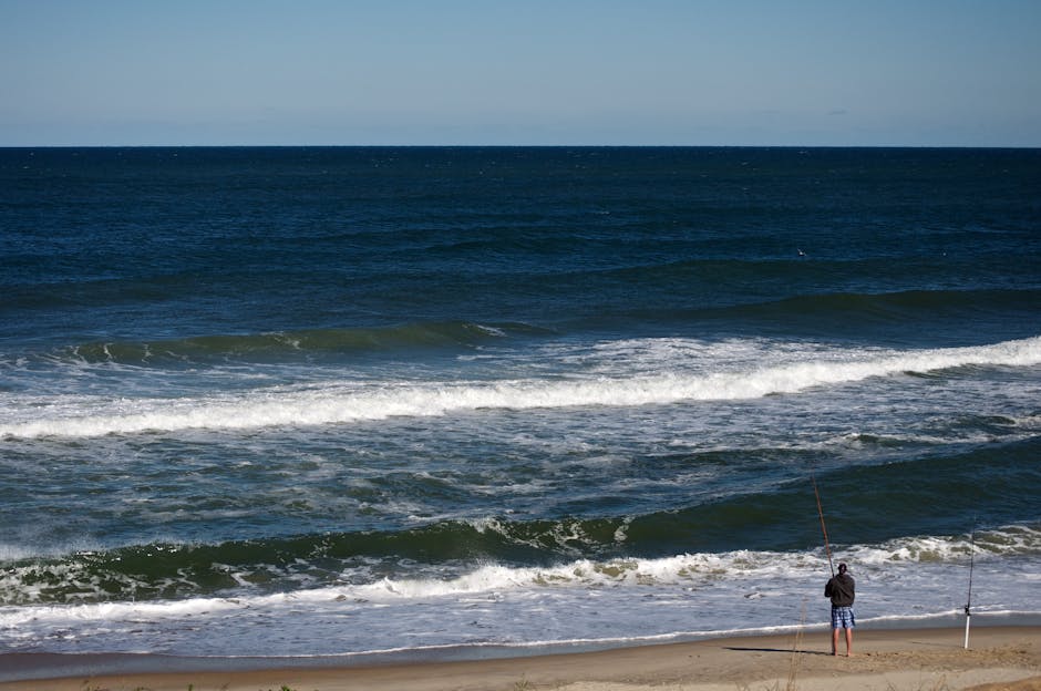 A man standing on a sandy beach near the shoreline, holding a fishing rod, appears to be preparing for or engaging in fishing as small waves roll onto the shore. The scene includes a light-colored sandy beach, with a gentle incline towards the water, and the ocean extending into the distance with layers of white-capped waves. The sky is clear with no visible clouds, and the water shows varying shades of blue and green, indicating different depths. Near the man, a slim metal pole is planted in the sand, possibly marking a fishing spot or boundary, and the man is dressed in casual clothing, including a dark jacket and shorts. The scene reflects a calm seascape, and the overall environment suggests a peaceful day by the coast, suitable for leisure activities like fishing and relaxing by the sea, relevant to home relocation or leisure-related services supported by Man with Van Nags Head.