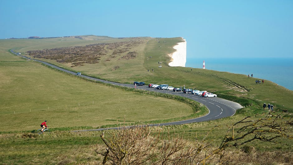 A wide view of the Seven Sisters cliffs on the south coast of England showing a winding coastal road with several parked cars along the side. The foreground features a cyclist riding on a grassy path, while a few pedestrians walk near the cliff edge on a paved pathway. In the background, the white chalk cliffs rise sharply above the ocean, with some visitors visible on the grassy slopes and walking along narrow trails. The landscape includes lush green fields, scattered bushes, and a distant lighthouse situated on the cliff's edge. Bright daylight illuminates the scene, capturing the natural beauty of the coastal scenery adjacent to the beach and chalk formations, suitable for content related to house removals or relocation logistics in the area associated with Man with Van Nags Head.