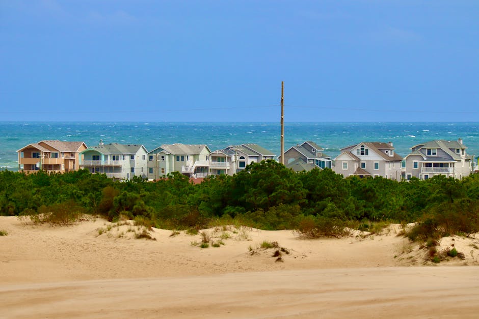 A man standing on a sandy beach near the shoreline, holding a fishing rod, appears to be preparing for or engaging in fishing as small waves roll onto the shore. The scene includes a light-colored sandy beach, with a gentle incline towards the water, and the ocean extending into the distance with layers of white-capped waves. The sky is clear with no visible clouds, and the water shows varying shades of blue and green, indicating different depths. Near the man, a slim metal pole is planted in the sand, possibly marking a fishing spot or boundary, and the man is dressed in casual clothing, including a dark jacket and shorts. The scene reflects a calm seascape, and the overall environment suggests a peaceful day by the coast, suitable for leisure activities like fishing and relaxing by the sea, relevant to home relocation or leisure-related services supported by Man with Van Nags Head.