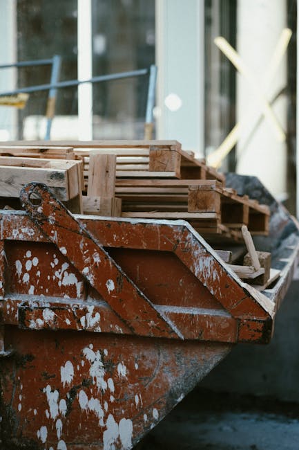 Close-up view of a rusted metal boat with visible white paint splatters, positioned outdoors near a building with large glass windows. Inside the boat, several packed wooden pallets and cardboard boxes with protective plastic wrap are stacked, prepared for transport during a home relocation or furniture transport service. The pallets and boxes are arranged on the boat's floor, ready for loading onto a vehicle, with some lifting straps and packaging materials nearby. In the background, a construction site or renovation area is partially visible with scaffolding and a partially completed wall. The scene captures the loading process typical of professional removals, highlighting equipment like trolleys and blankets used for moving bulky items efficiently and safely. The environment is well-lit, emphasizing the detailed textures of the pallets, cardboard, and the boat’s surface, aligning with the context of packing and moving operations associated with house removals near Nags Head.
