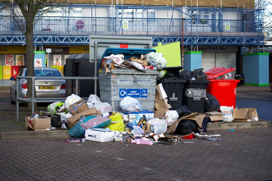 Photograph of multiple overflowing waste collection bins and surrounding rubbish outside a commercial building on a city street during daytime. The image shows large grey, black, and red bins with open lids, filled with mixed paper, cardboard, plastic bags, and miscellaneous household refuse. Loose items, including cardboard boxes, plastic packaging, and paper waste, are scattered on the ground around the bins, with some materials spilling onto the paved pavement and sidewalk. The scene is positioned in front of a building with storefronts, with a metal railing separating the waste area from parked vehicles, including a silver van. The background includes a multi-storey building with scaffolding and a sign indicating a fish bar. The image depicts typical home relocation or removal logistics, highlighting the importance of proper disposal practices, and is associated with services like those offered by Man with Van Nags Head.