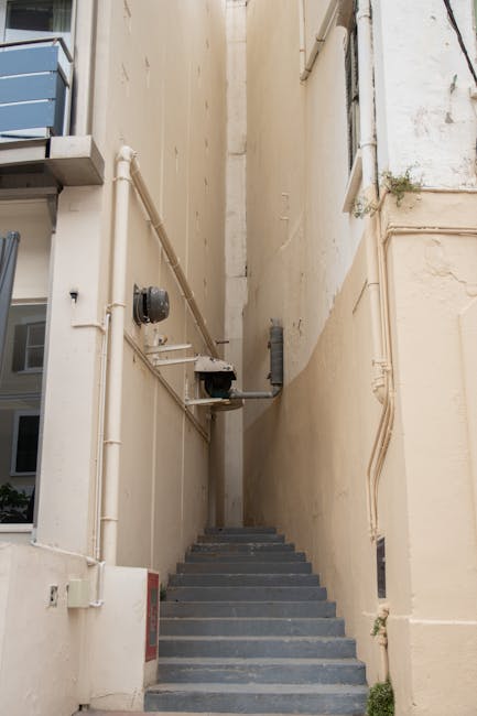 A narrow outdoor stairwell situated between two beige-painted building walls, with a series of grey concrete steps leading upward. To the left, there are exposed utility pipes, including a black meter and white PVC pipes, along with an outdoor electrical box mounted on the wall. To the right, small plants are growing from cracks in the wall. The lighting appears natural, indicating daytime. This stairwell presents a logistical challenge for house removals where furniture and packing materials need to be carried through a confined space, as part of the loading process involved in home relocation in the Nags Head area. Man with Van Nags Head may need to navigate this tight space when transporting items to or from the property to ensure safe and efficient removal service.
