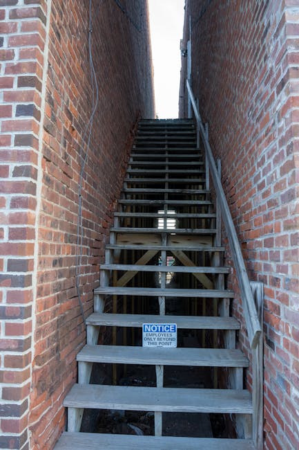 A narrow outdoor staircase constructed of unfinished wooden steps with a simple handrail on the right side, situated between two red brick walls. The staircase appears to lead up to a confined outdoor space or entrance area, with a small notice sign attached to the bottom step indicating restricted access beyond this point. The surrounding environment suggests a tight tower or alleyway, which can pose challenges during home relocation or furniture transport. The scene is illuminated by natural daylight, highlighting the utilitarian nature of the structure. This image exemplifies the type of staircase that Man with Van Nags Head may encounter when managing removals in constrained urban environments around Nags Head, where careful planning is required for safe and efficient loading or packing and moving operations.