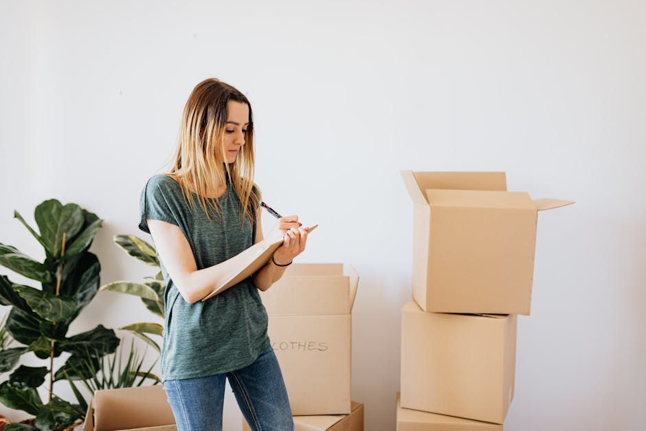 A young woman with long blonde hair, dressed in a casual grey t-shirt and jeans, stands inside a property surrounded by several cardboard moving boxes in various stages of being packed or unpacked. She is holding a pen and appears to be making a checklist or writing notes on a notepad. The boxes are labeled, with one clearly marked 'CLOTHES,' indicating packing for a home relocation. Behind her, a large green potted plant adds a touch of interior décor, and the room has bright, natural lighting with a plain white wall as the background. The scene captures the process of packing and organizing items for a house move, representing clean, organized packing for a professional removal service such as Man with Van Nags Head, which specializes in house removals and furniture transport during moves. The image emphasizes careful planning and documentation involved in a professional moving or packing process, aligned with the context of the tenant move checklist for Holloway Road, Nags Head.