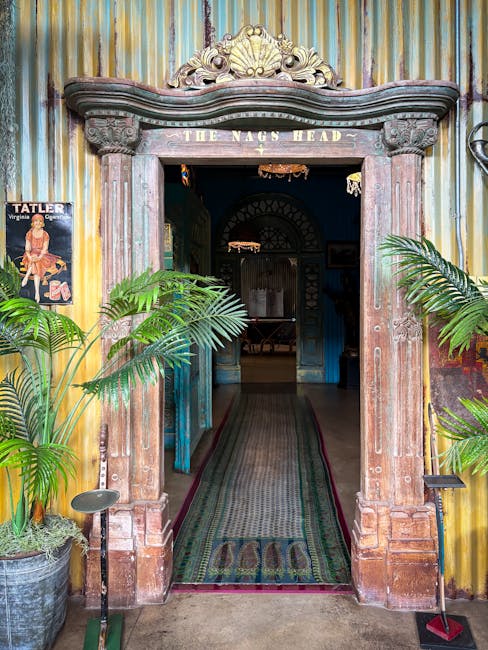 The image shows an ornate wooden doorway with decorative carvings and the inscription 'THE NAGS HEAD' above the entrance, leading into a dimly lit interior space. Flanking the entrance are large potted green plants, with one on each side, placed on the floor. The exterior wall features colorful, vertically striped metal panels. Inside, a narrow corridor with a patterned rug running along its length is visible, leading to a space beyond with arched features. The setting suggests a traditional pub or historic building exterior, relevant to house and business relocations, with natural lighting highlighting the details of the doorway and plants.